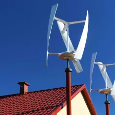Dwa białe, pionowe turbiny wiatrowe obracają się na tle błękitnego nieba, nad czerwonym dachem domu.