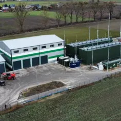 Nowoczesna biometanownia z zielonym dachem i kopułą, otoczona polami i budynkami gospodarczymi.