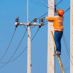 Elektryk w pomarańczowej kamizelce i kasku wspina się po drabinie, by naprawić linie energetyczne. Praca przy wysokim napięciu wymaga precyzji i doświadczenia, by zapewnić bezpieczeństwo i ciągłość dostaw.