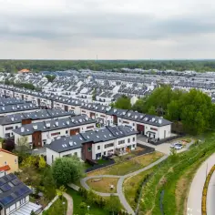 Widok z lotu ptaka na osiedle domów jednorodzinnych z ciemnymi dachami i białymi elewacjami, w pobliżu drogi.