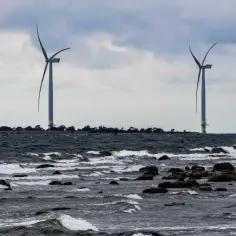 Dwie turbiny wiatrowe na morzu, symbol nowoczesnych farm wiatrowych w Polsce. Woda faluje, kamienie wystają z morza.
