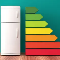 Biała lodówka obok kolorowej drabinki klas energetycznych, gdzie zielony oznacza najlepszą, a czerwony najgorszą. Ta lodówka ma klasę energetyczną E.