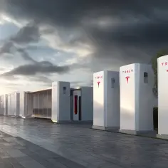 Rząd białych, pionowych jednostek magazynujących energię z logo Tesli, stojących na utwardzonym podwórku. Słońce zachodzi za chmurami.