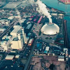 Ogromna spalarnia śmieci z charakterystyczną kopułą i kominami, z których unosi się dym. Wokół kompleks przemysłowy i port.