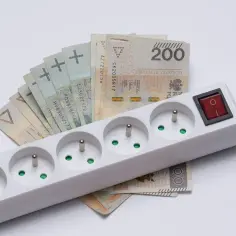 Biała listwa z gniazdkami i czerwonym włącznikiem, pod którą leżą polskie banknoty. Symbolizuje dopłaty do prądu.