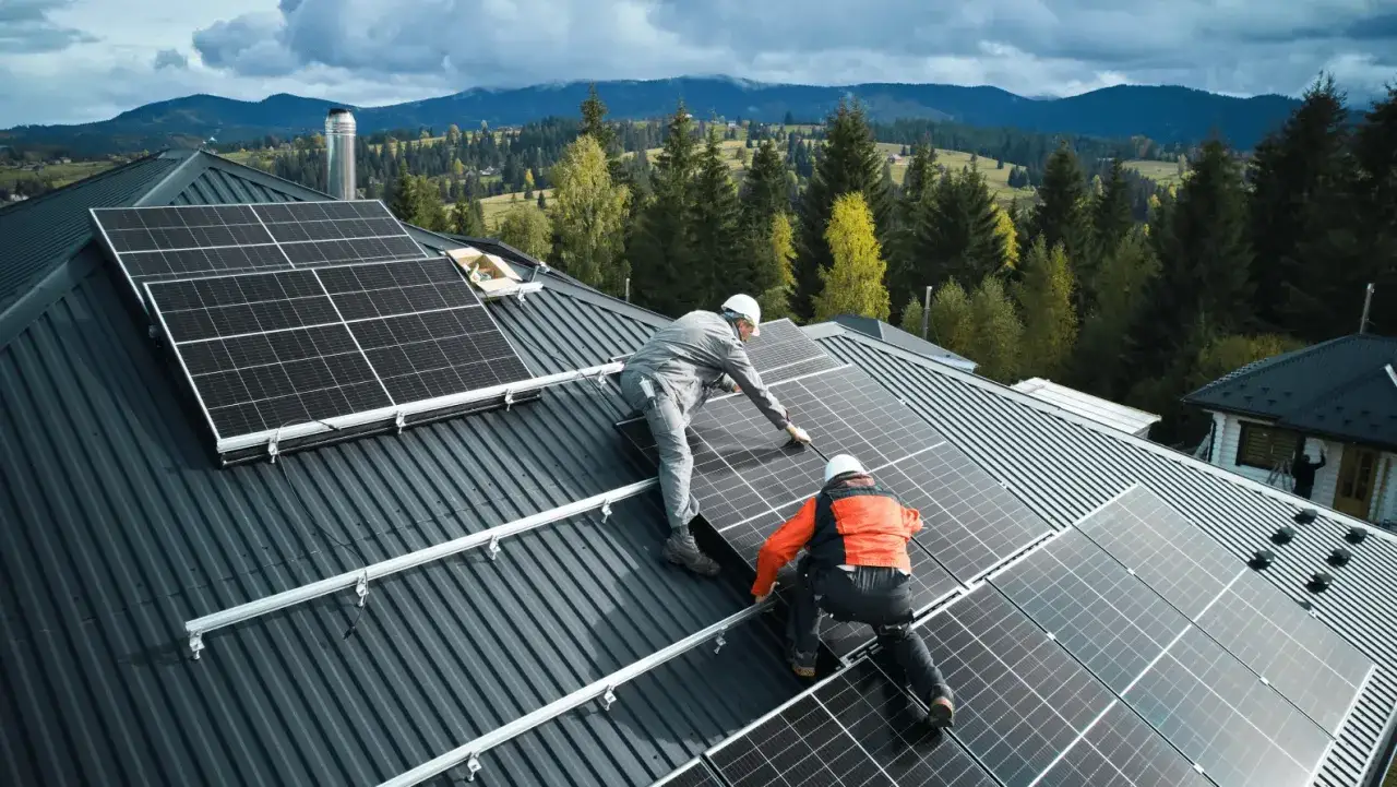 Instalacja fotowoltaiczna 10 kw cena z montażem. Dwóch monterów na dachu instaluje panele słoneczne. W tle góry i las.